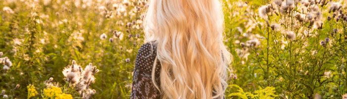 Domowe sposoby na alergię na pyłki. Atrakcyjna, młoda dziewczyna o długich blond włosach stoi na łące, zwrócona tyłem do aparatu.