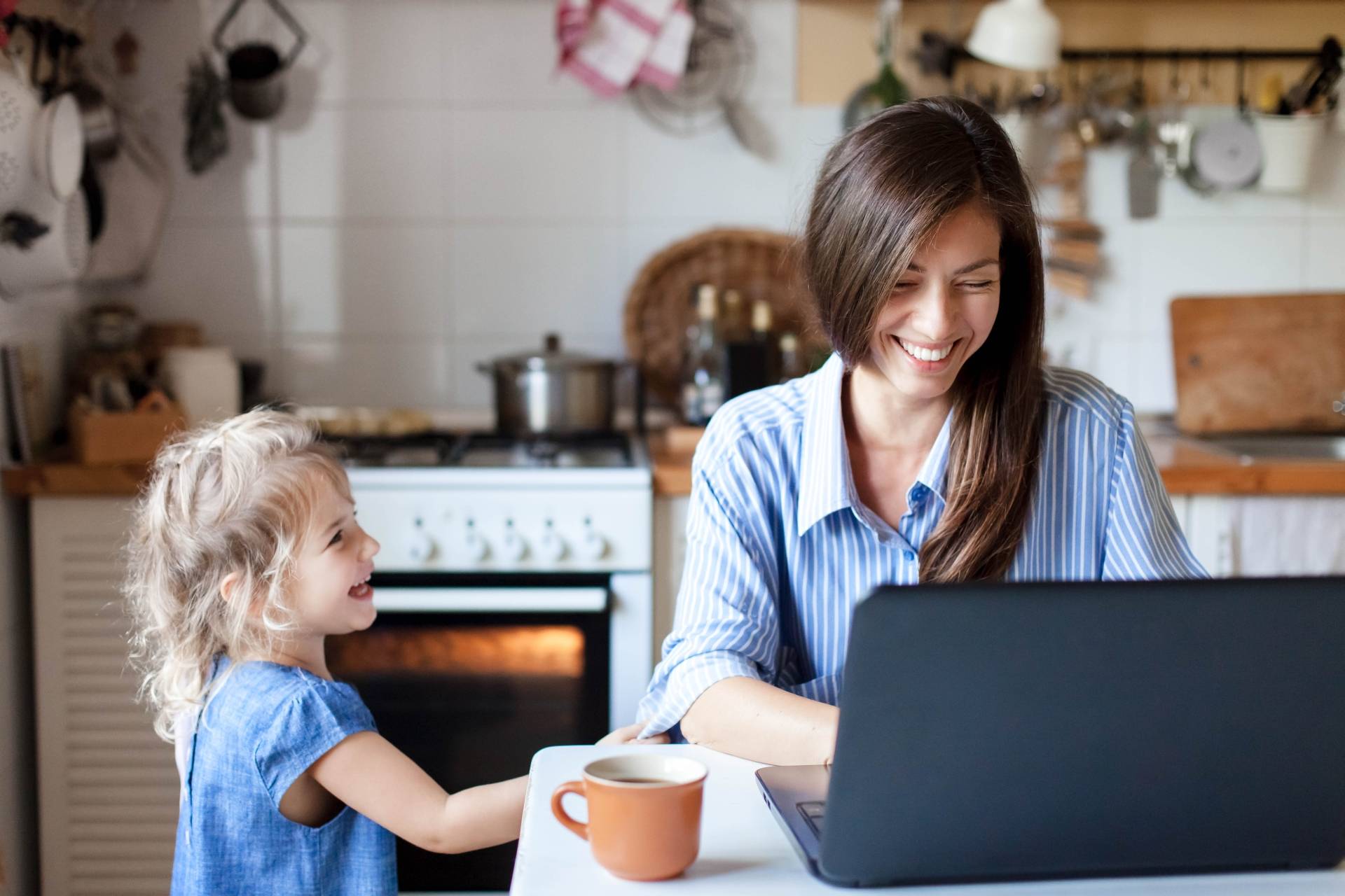 Work-life balance w dobie home office. Mama pracuje na laptopie w kuchni, obok stoi kilkuletnia córeczka i zachęca ją do zabawy.