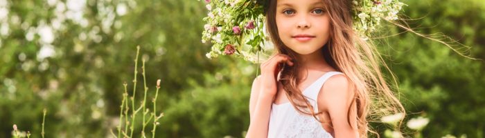 Dziewczynka o długich blond włosach w białej letniej sukience i pięknym wielkim wianku z kwiatów na głowie uśmiecha się do obiektywu. Zioła Matki Boskiej Zielnej.