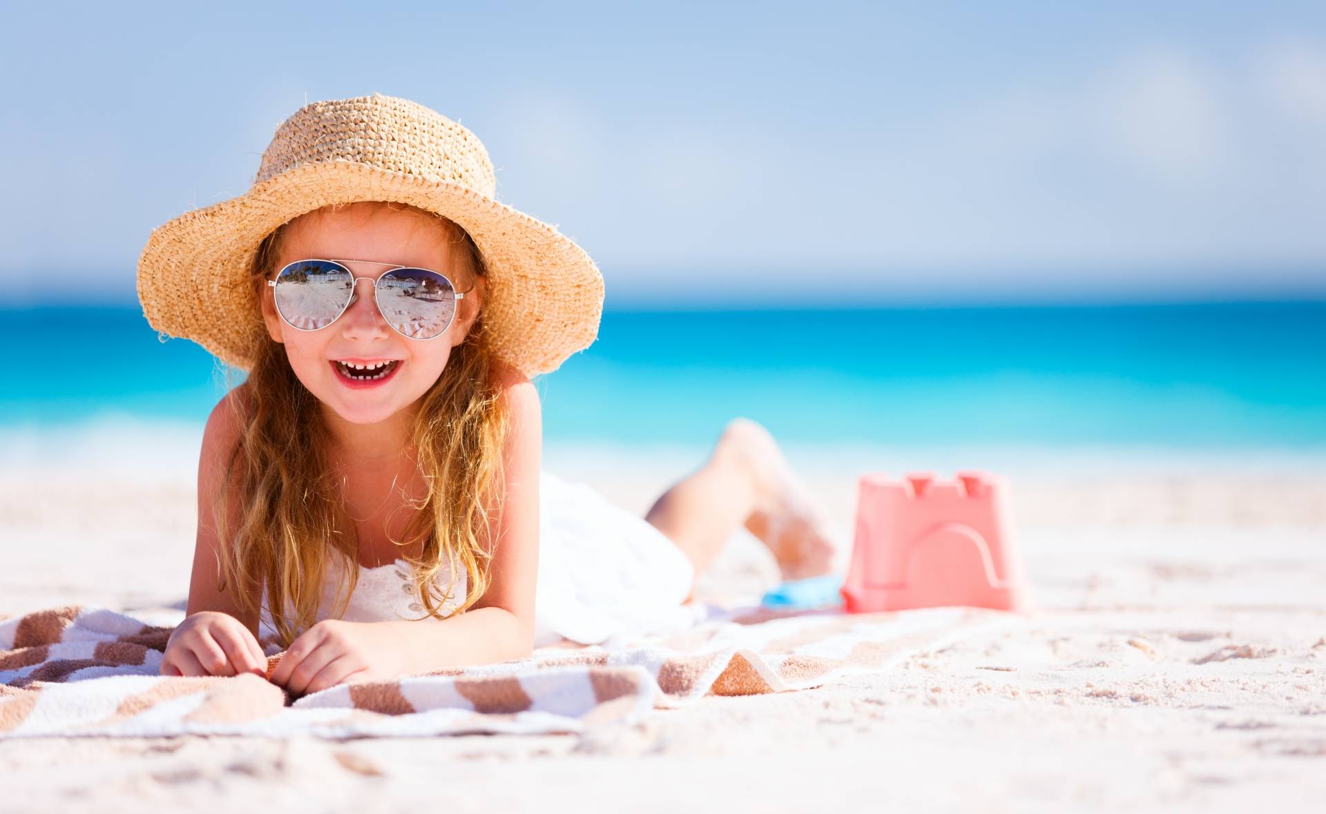 Dziewczynka w okularach przeciwsłonecznych i kapeluszu słomkowym opala się na plaży. Jak chronić dziecko przed udarem słonecznym? 