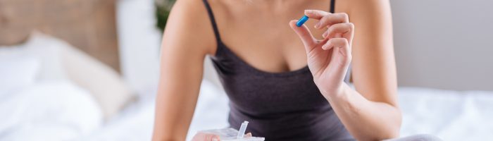 Efekt nocebo - co to jest i na czym polega? Kobieta w szarym dresie siedzi na łóżku po turecku i przygląda się podejrzliwie niebieskiej pigułce, którą ma połknąć.