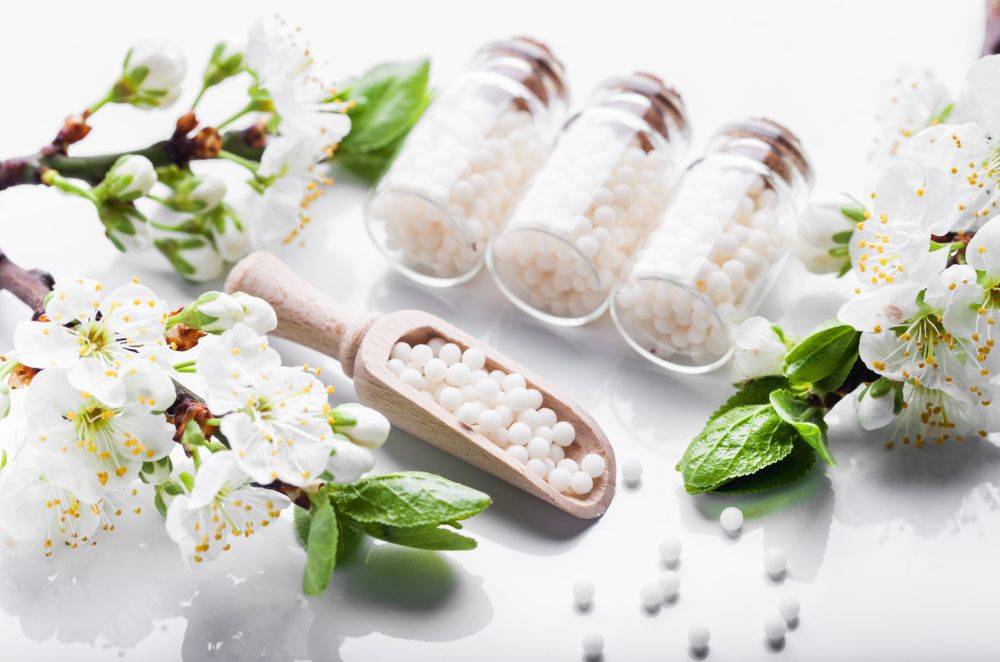 Homeopatia - leki homeopatyczne w słoiczkach ułożone na białym blacie w otoczeniu białych kwiatów jaśminu.