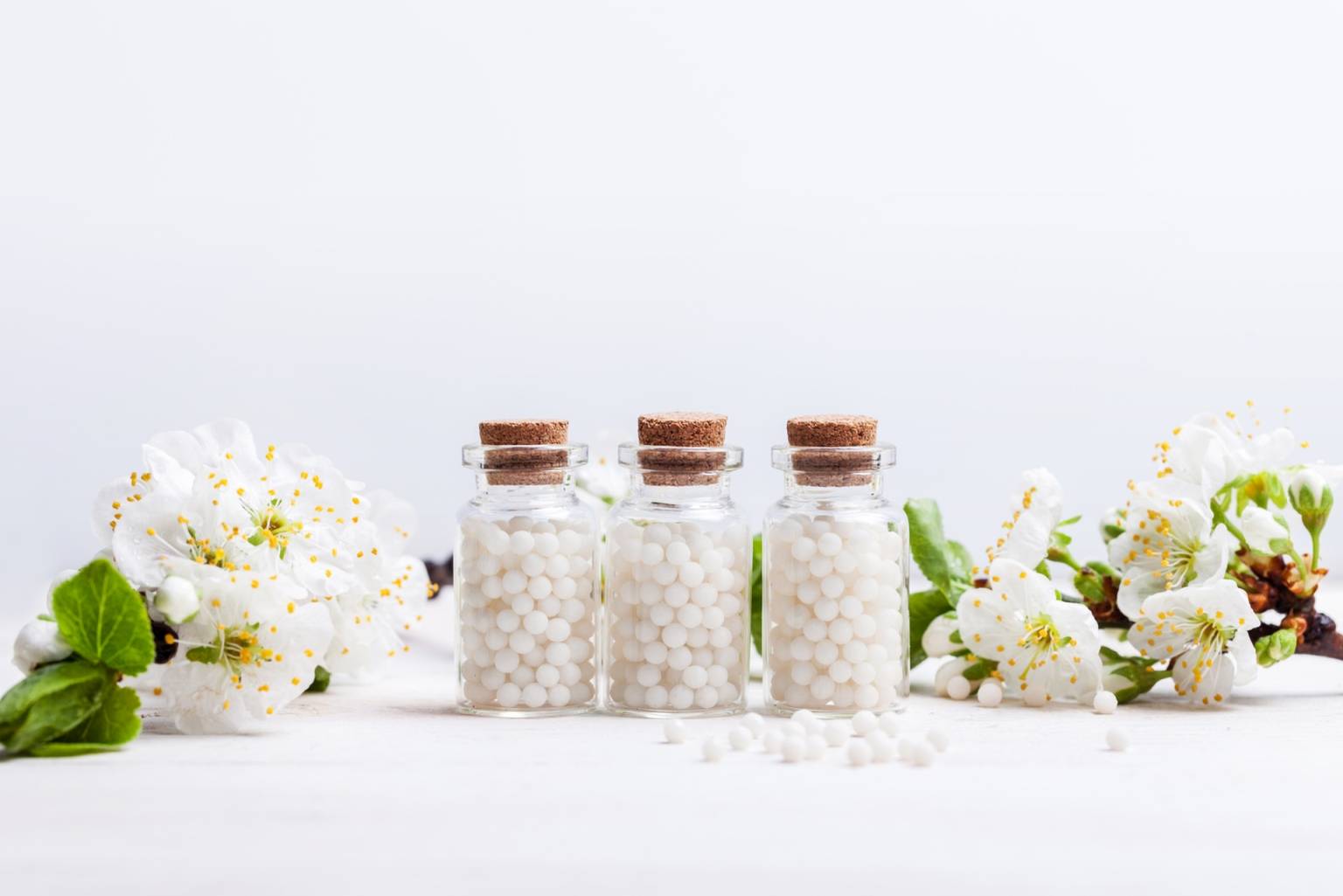 Leki homeopatyczne na objawy rotawirusa u dzieci. Jak pokonać grypę żołądkową u dziecka? Trzy fiolki z granulkami homeopatycznymi stoją na białym blacie w otoczeniu kwiatów jabłoni.