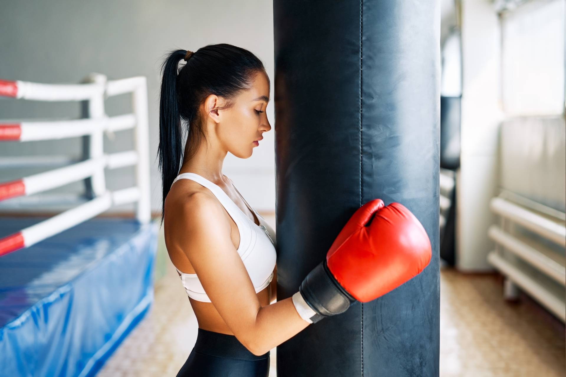 Myśl jak sportowiec - jak czerpać mądrość z psychologii sportu? Młoda atrakcyjna kickbokserka opiera się głową o worek treningowy i obejmuje go rękawicami.