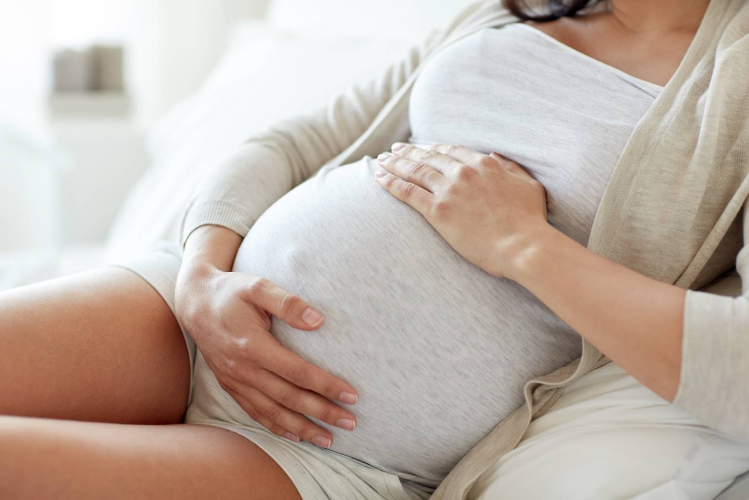 Leczenie niepłodności w medycynie chińskiej. Kobieta w zaawansowanej ciąży w szarej bluzce, narzutce i krótkich spodenkach siedzi na kanapie i trzyma się za brzuch.