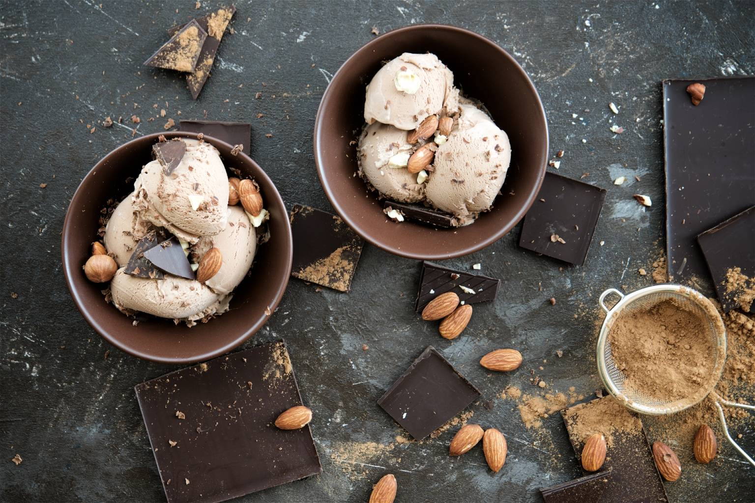Lody naturalne domowe a'la snickers w miseczkach, obok leżą migdały i kawałki gorzkiej czekolady.