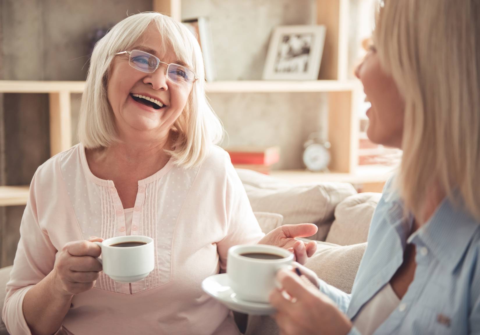 Relacje matka i córka - jak o nie zadbać? Mama seniorka i dorosła córka siedzą w salonie, piją kawę i się śmieją.