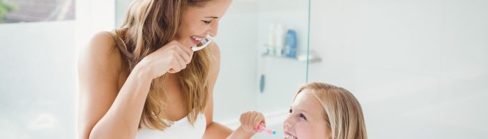 Jak dbać o zdrowe i mocne zęby i higienę jamy ustnej? Mama i córeczka ubrane w białe koszulki myją zęby w łazience.