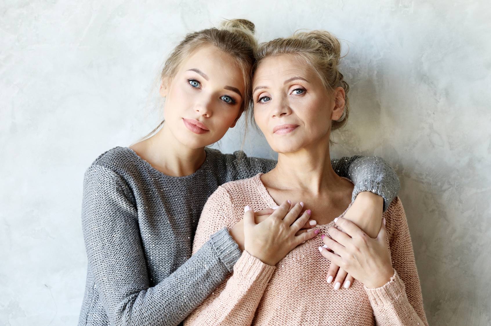 Relacje matka i córka - gdy dziecko się usamodzielnia. Mama i dorosła córka stoją na tle szarej ściany, przytulają się do siebie i patrzą w aparat.
