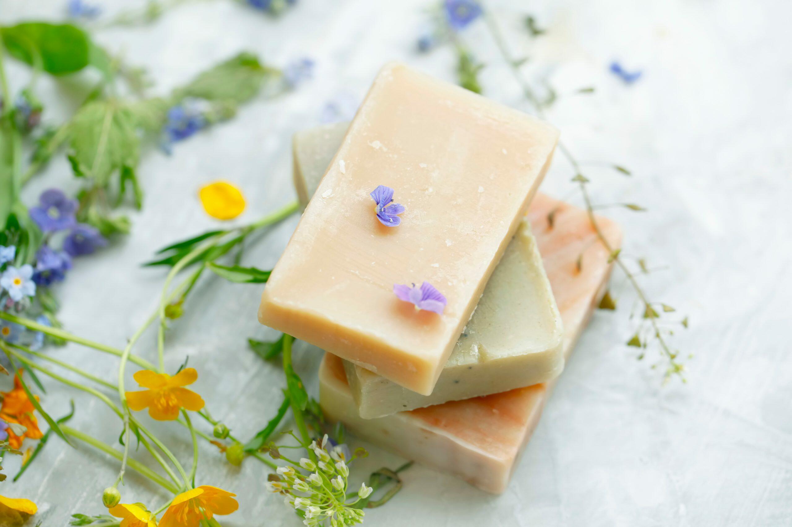 Kosmetyki w kostce - jak używać szamponu w kostce i jak zrobić własny balsam w kostce DIY? Kostki naturalnego mydła ułożone na sobie w otoczeniu kwiatów i zielonych liści.