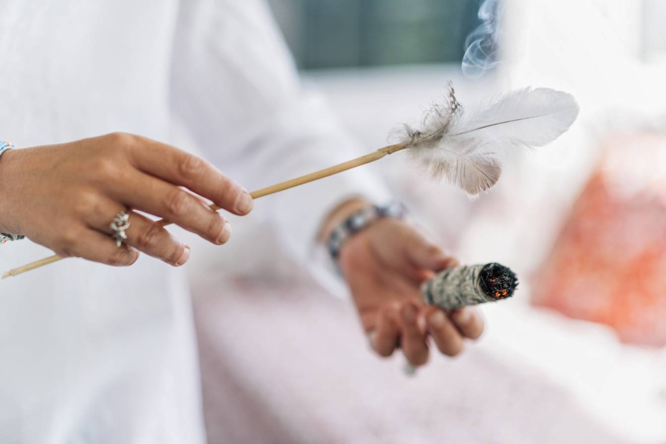 Okadzanie domu palo santo i białą szałwią - czy jest zdrowe i ekologiczne? Kobieta w białej koszuli boho okadza dom za pomocą kadzidła i piórka do rozprzestrzeniania dymu.
