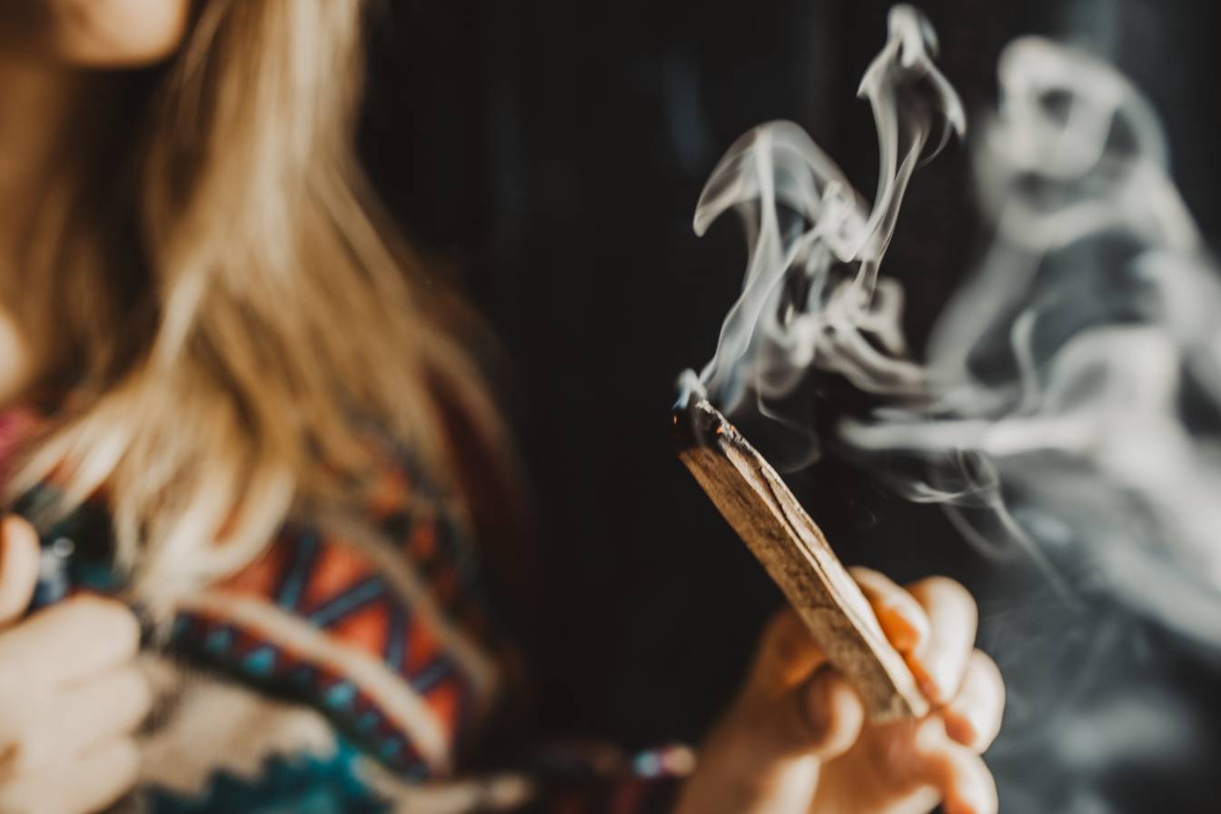 Jakie właściwości ma palo santo i biała szałwia do okadzania domu? Kobieta w etnicznym ponczo okadza pomieszczenie palo santo. Ciemne tło.