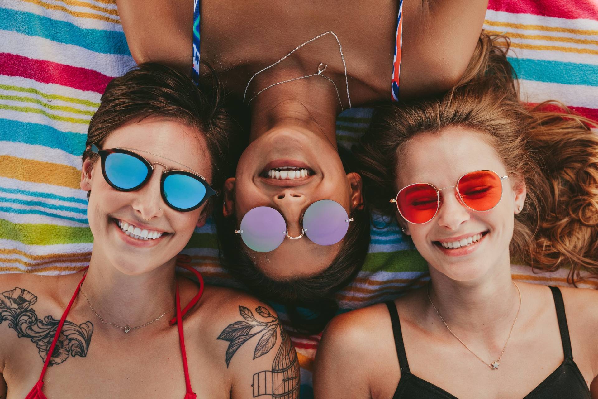 Jak się bezpiecznie opalać przy problemach z cerą? Trzy przyjaciółki w okularach przeciwsłonecznych opalają się na plaży.
