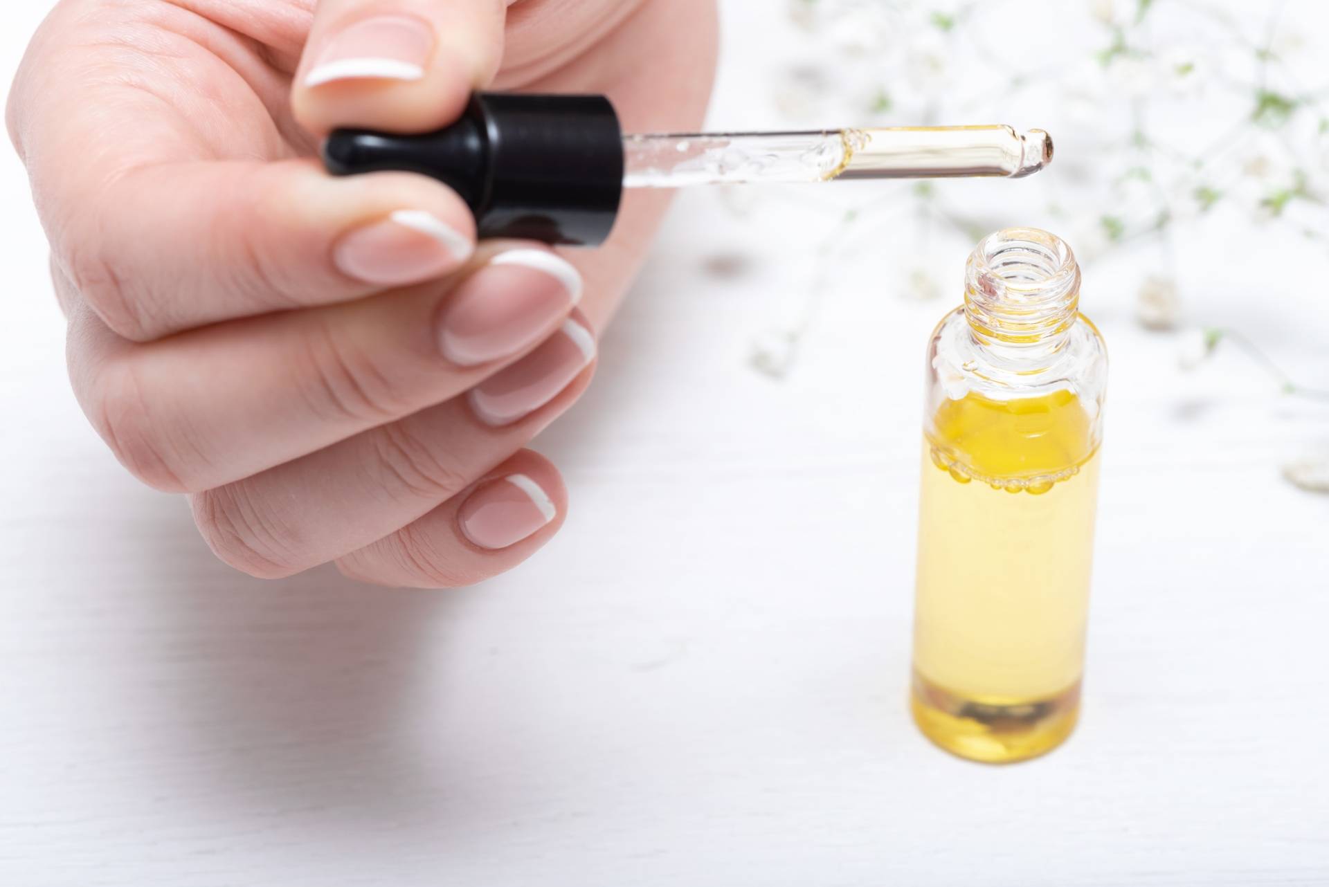 Olejek arganowy do pielęgnacji paznokci. Kobieca dłoń z wypielęgnowanymi paznokciami trzyma pipetę nad buteleczką olejku stojącą na białym blacie. W tle białe kwiaty.