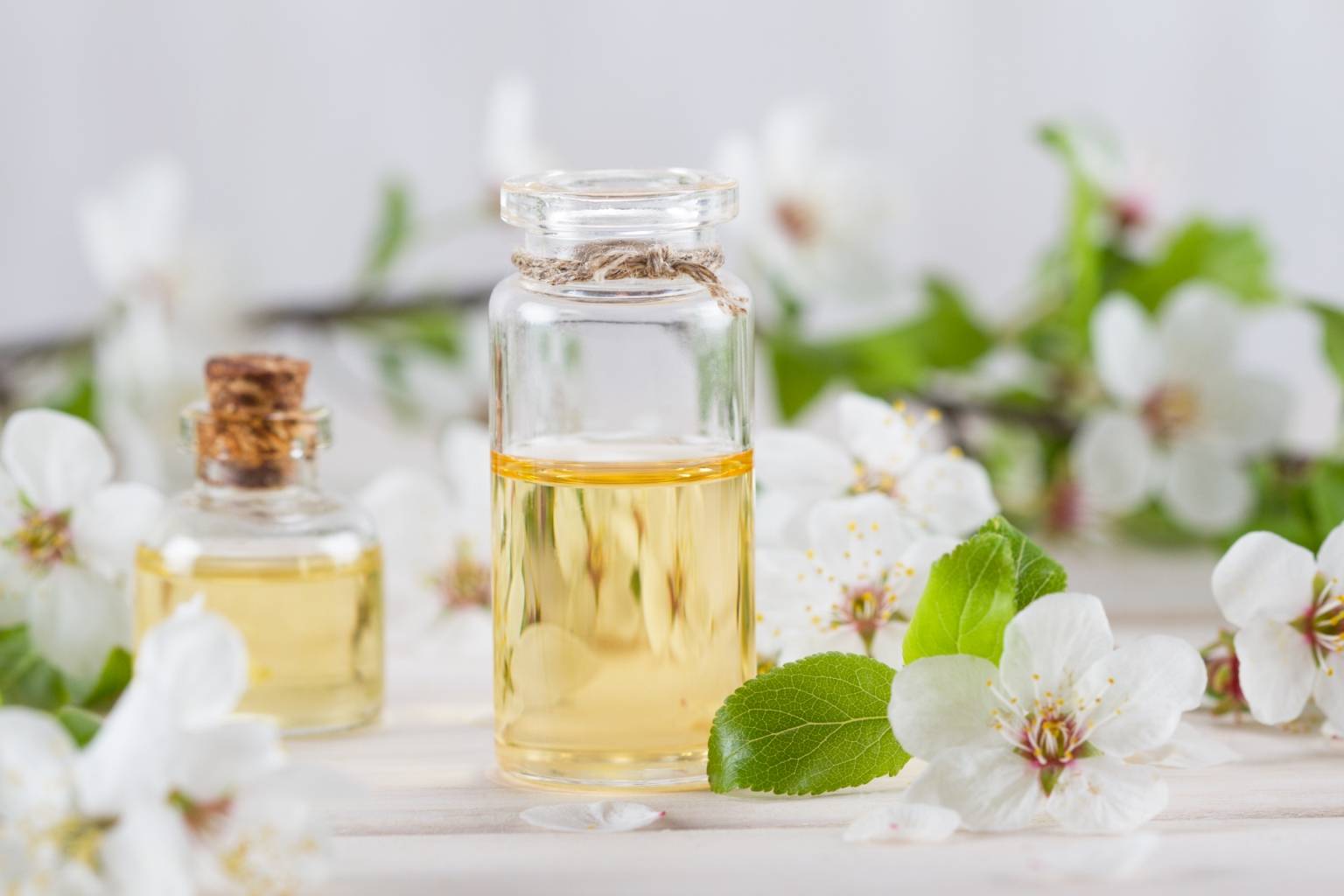 Czym są olejki eteryczne i jak działają? Na czym polega aromaterapia? Jasnożółty olejek w przezroczystej buteleczce stoi na jasnym blacie w otoczeniu białych kwiatów jabłoni.