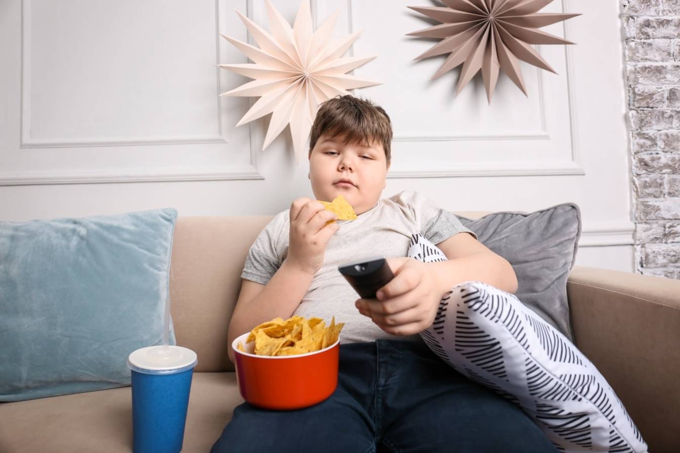 Otyłość u dzieci - jak z nią walczyć? Chłopiec z dużą nadwagą siedzi na kanapie, je chipsy i ogląda telewizję.