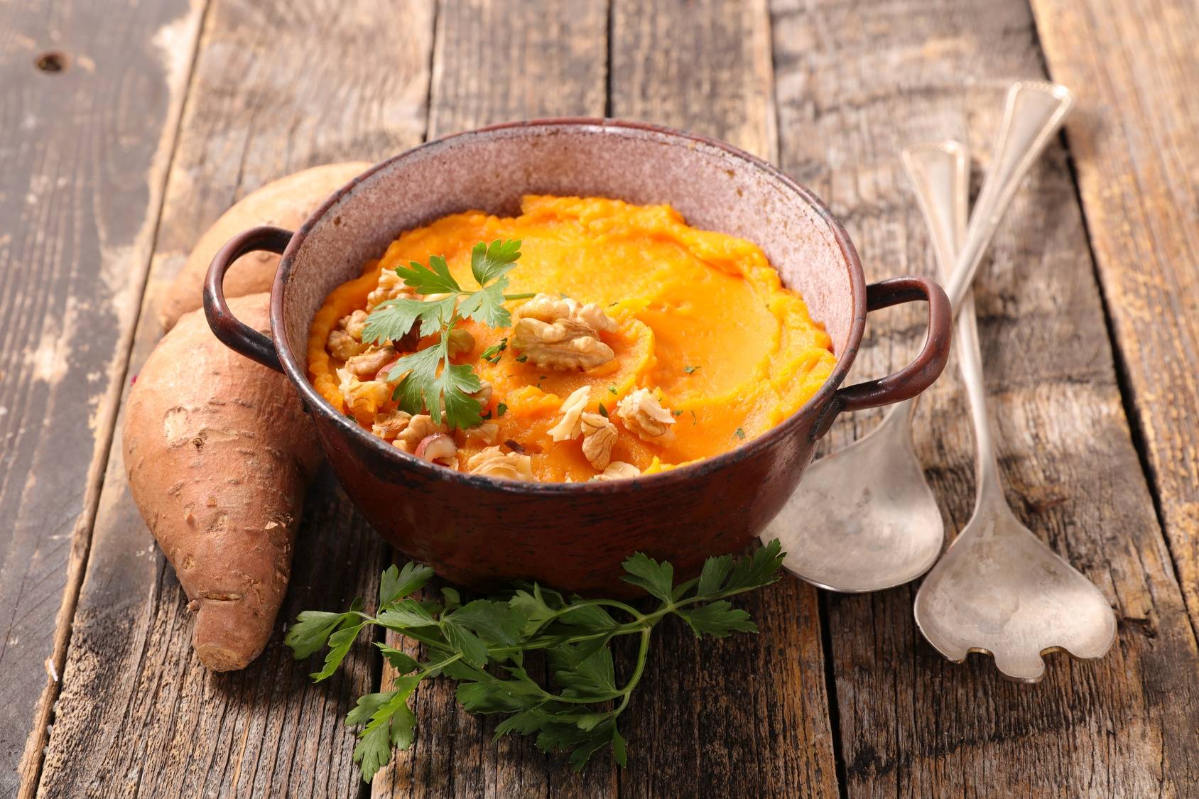 Puree z batatów w garnuszku żaroodpornym na drewnianym blacie, obok leży batat i sztućce - przepisy na dania z batatów.