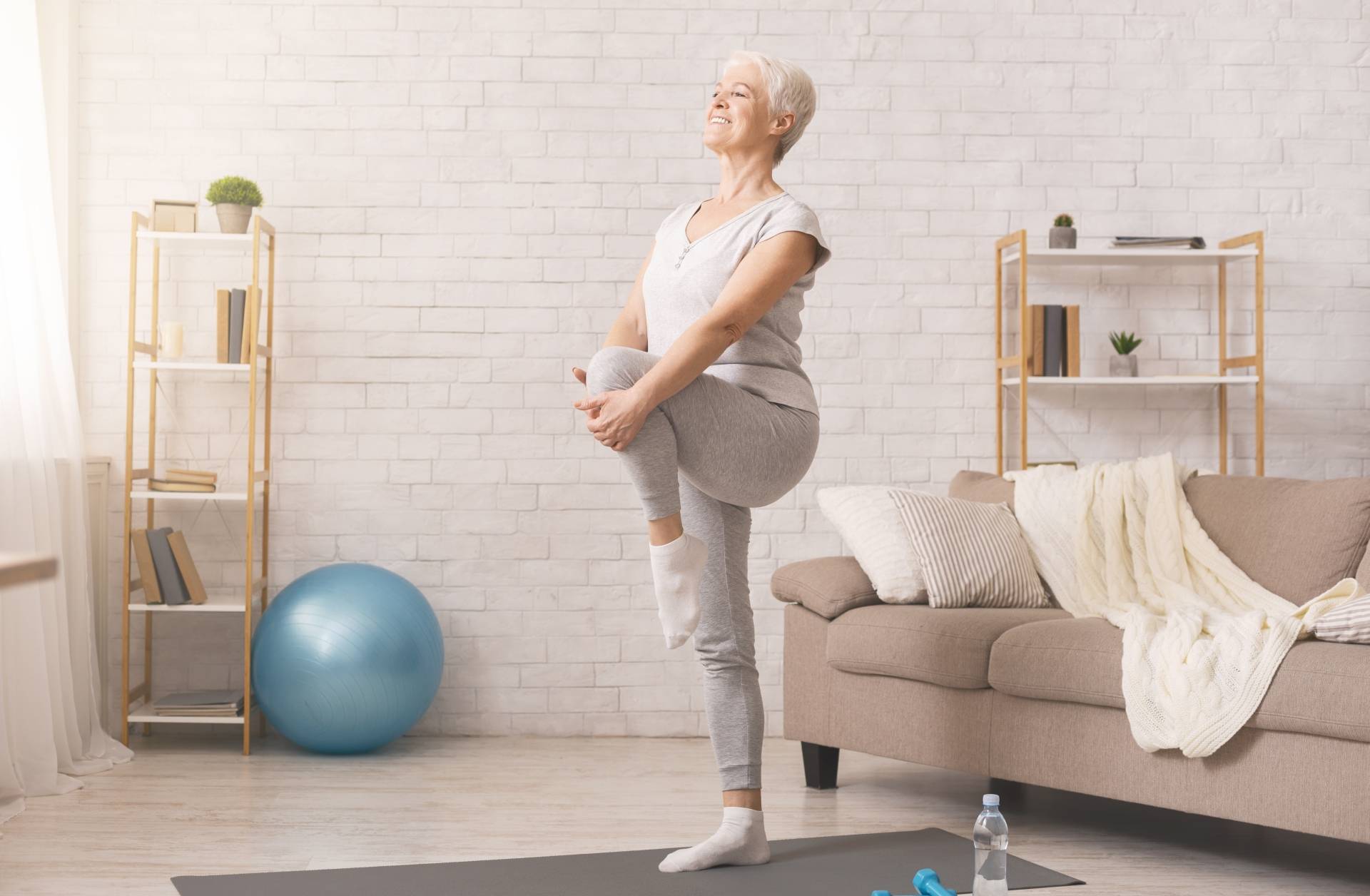Reumatyzm - objawy i leczenie. Aktywna seniorka w stroju sportowym gimnastykuje się na macie w domu, stoi z podniesioną nogą i przyciąga kolano do siebie.