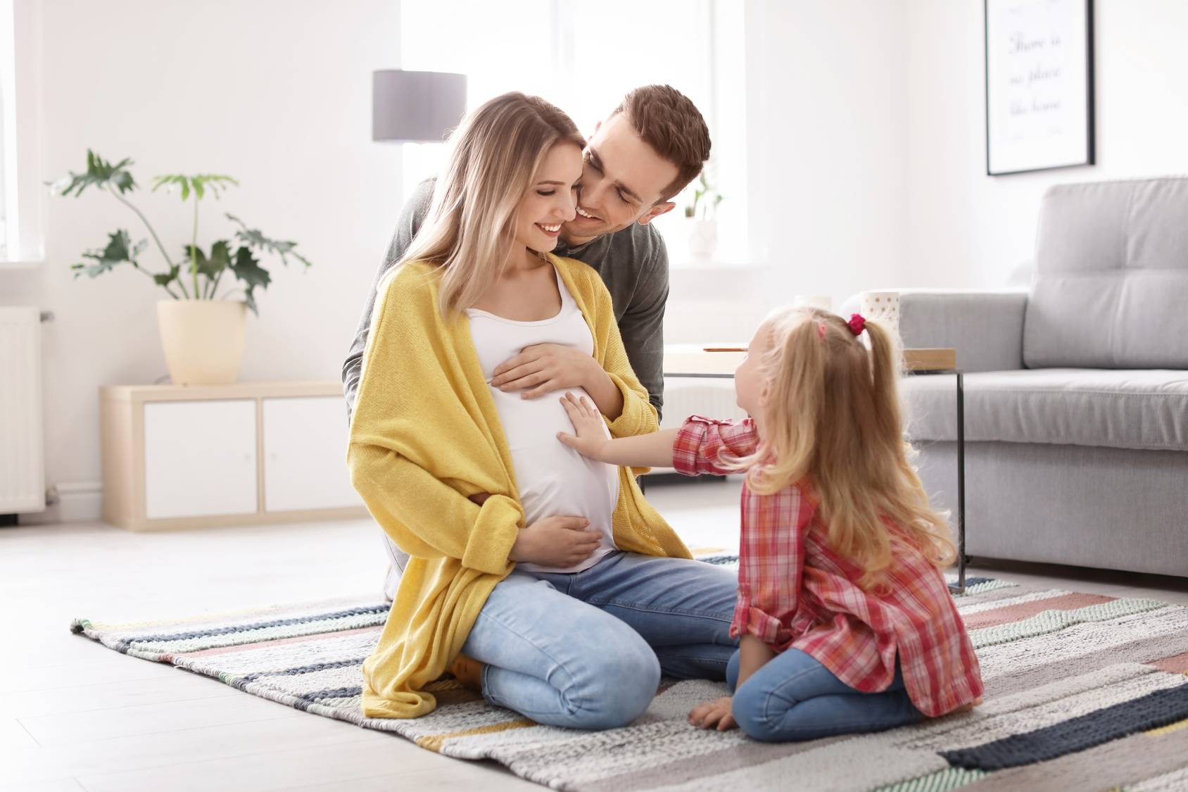 Jak zadbać o siebie w ciąży według tradycyjnej medycyny chińskiej? Kobieta w ciąży siedzi na dywanie w salonie wraz z rodziną - mężem i córeczką.