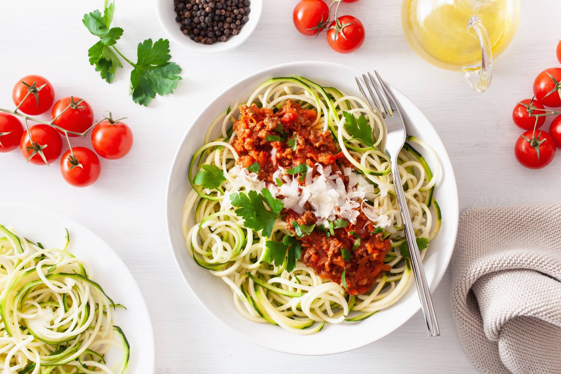 Spaghetti z cukinii na białym talerzu i białym blacie. Obok leżą pomidorki koktajlowe, oliwa, miseczka z ziarnami pieprzu.