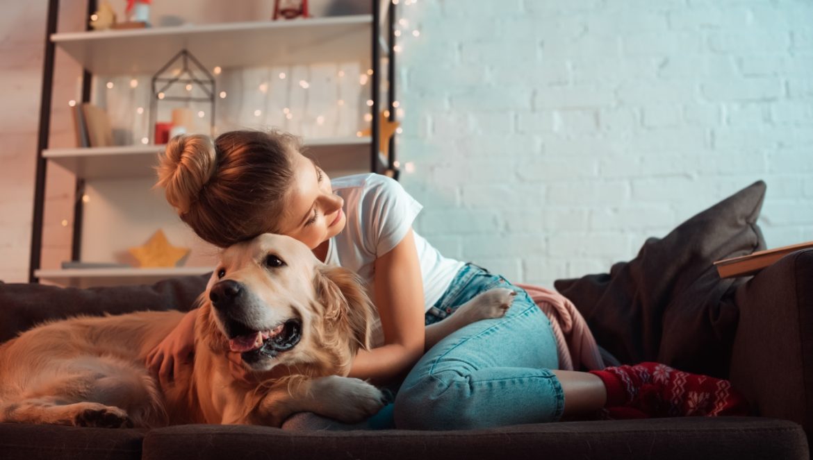 Miłość singielki - dobre życie bez partnera. Wywiad z psycholog Joanną Kot. Młoda kobieta w dżinsach i koszulce przytula się do swojego psa golden retrivera, siedząc na kanapie w domu.
