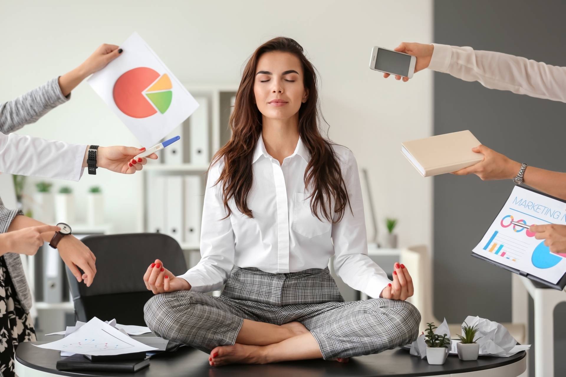 Work-life balance - jak go odnaleźć? Młoda kobieta, pracowniczka korporacji, ubrana w białą koszulę i szare materiałowe spodnie siedzi na biurku, medytuje. Pozostali pracownicy wyciągają w jej stronę ręce z notatkami i rzeczami do zrobienia.