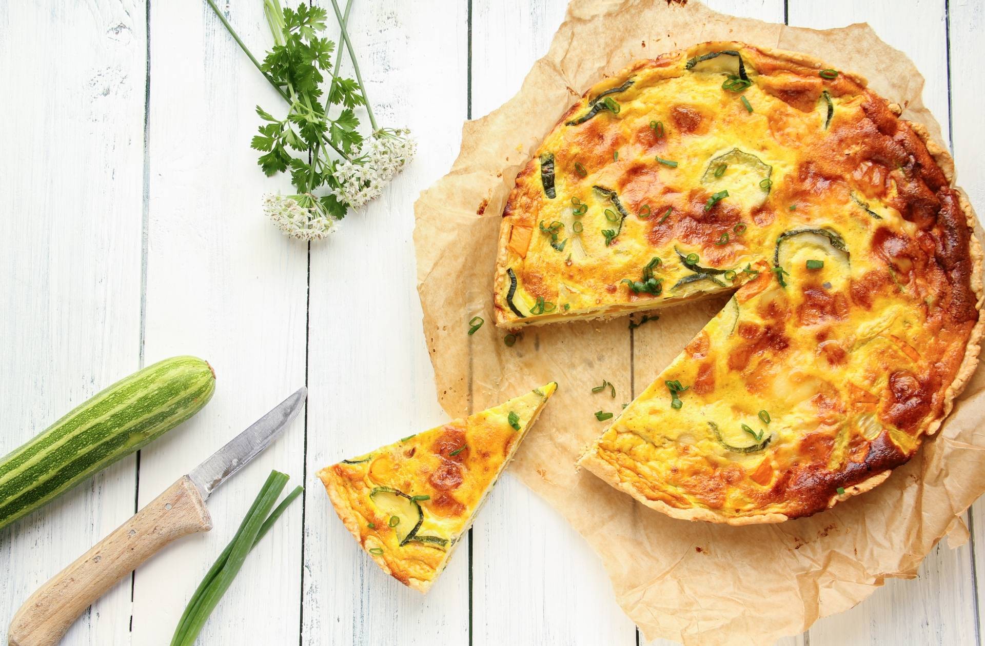 Tarta z cukinią na papierze do pieczenia na białym drewnianym blacie. Obok leży cukinia i nóż z drewnianą rączką.