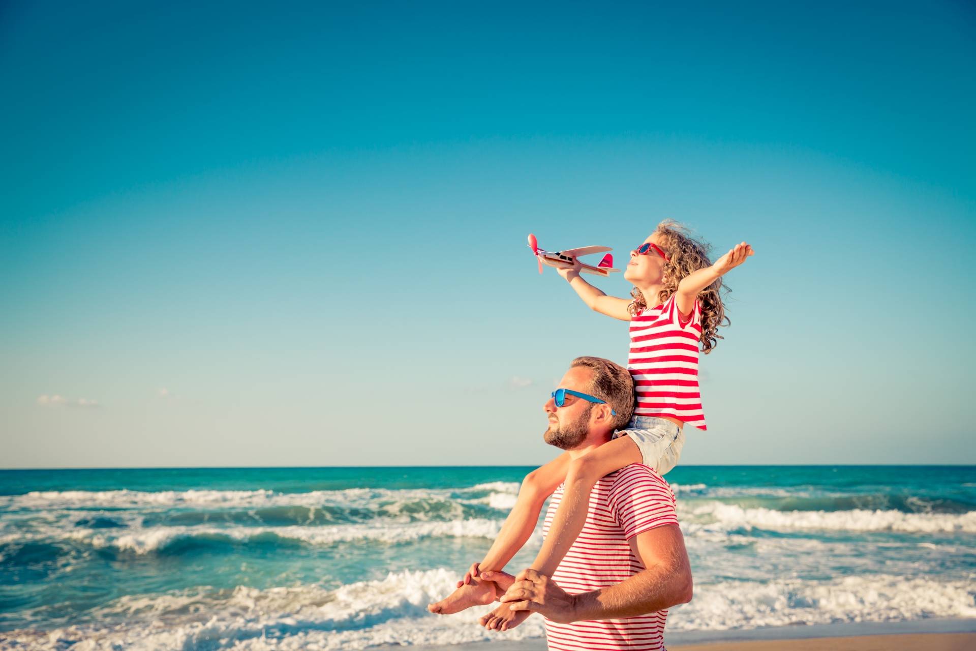 Dziewczynka siedzi u taty na barana i bawi się małym samolotem na plaży. Jak chronić dziecko przed udarem słonecznym?