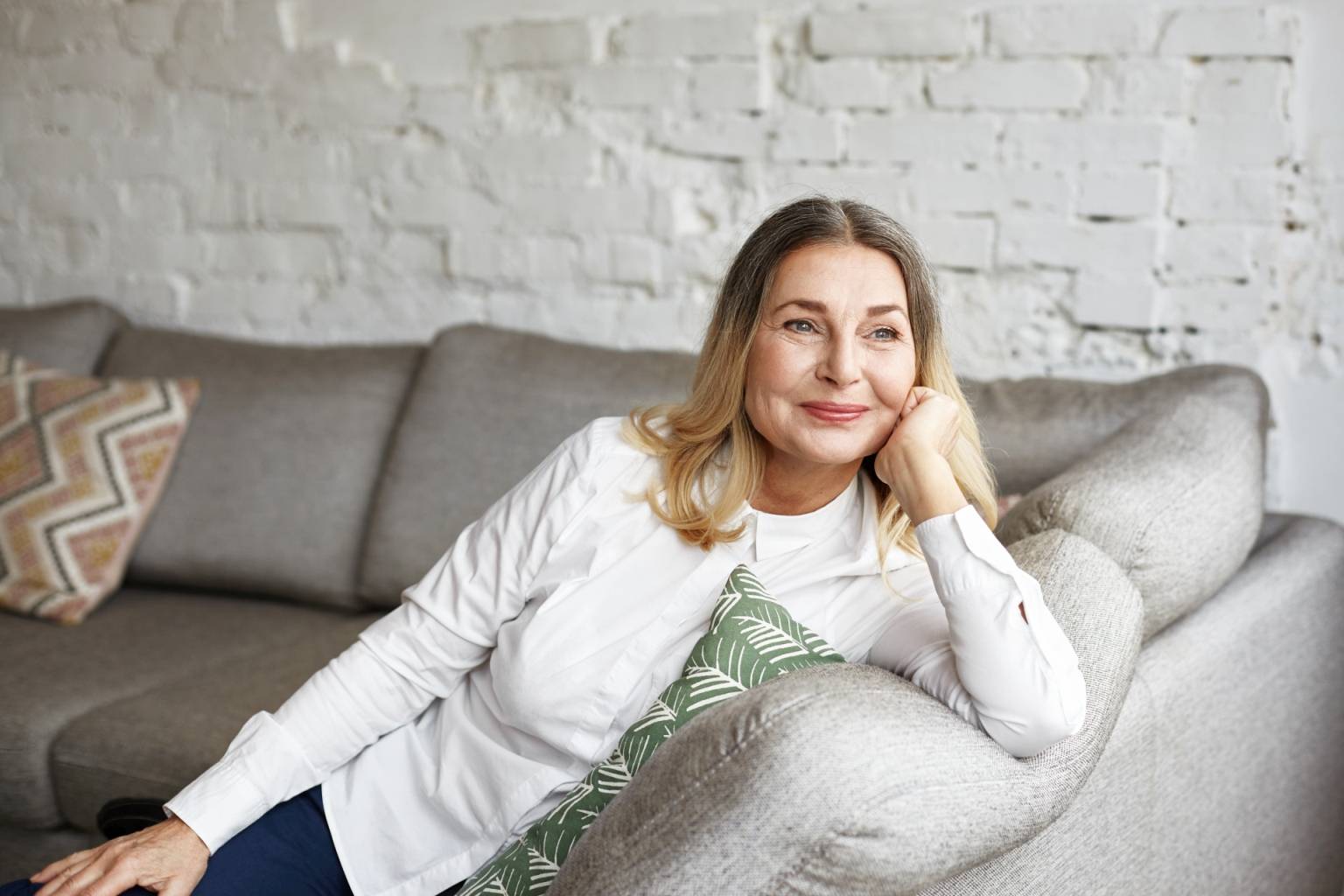 Leki homeopatyczne na menopauzę. Uśmiechnięta seniorka siedzi na kanapie w domu i wygląda przez okno.