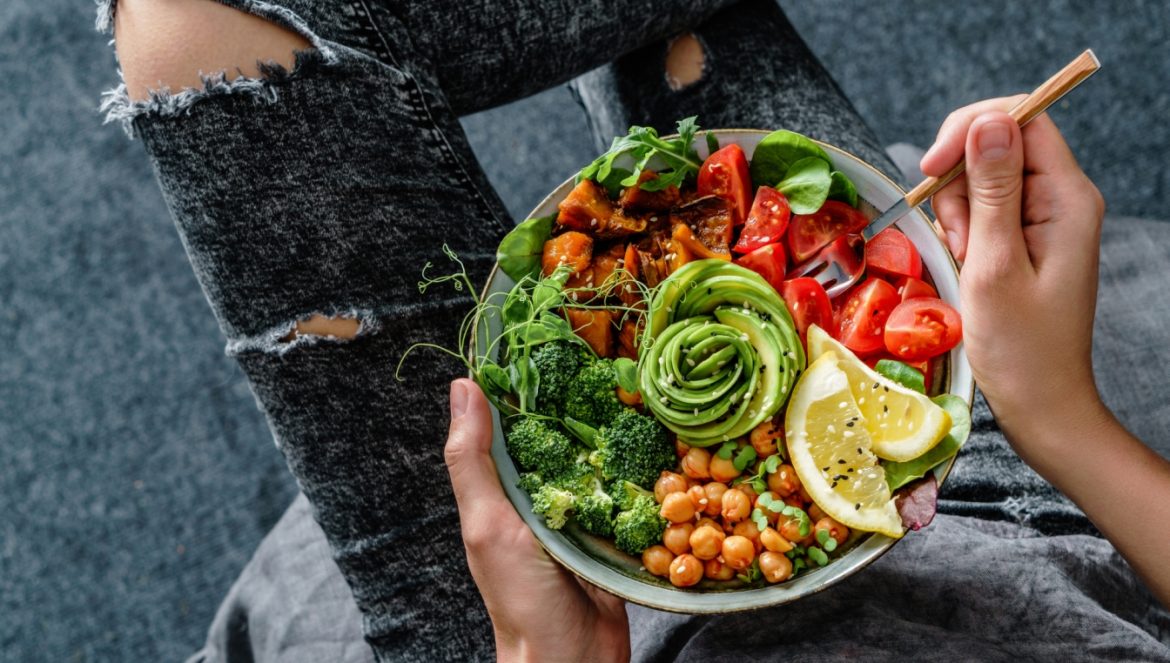 Buddha Bowl - jak skomponować miskę Buddy? Młoda kobieta hipsterka w czarnych przetartych jeansach z dziurami i szarym t-shircie trzyma w rękach michę obfitości z sałatą, pieczonymi batatami,, cieciorką, brokułami, awokado i fasolką.