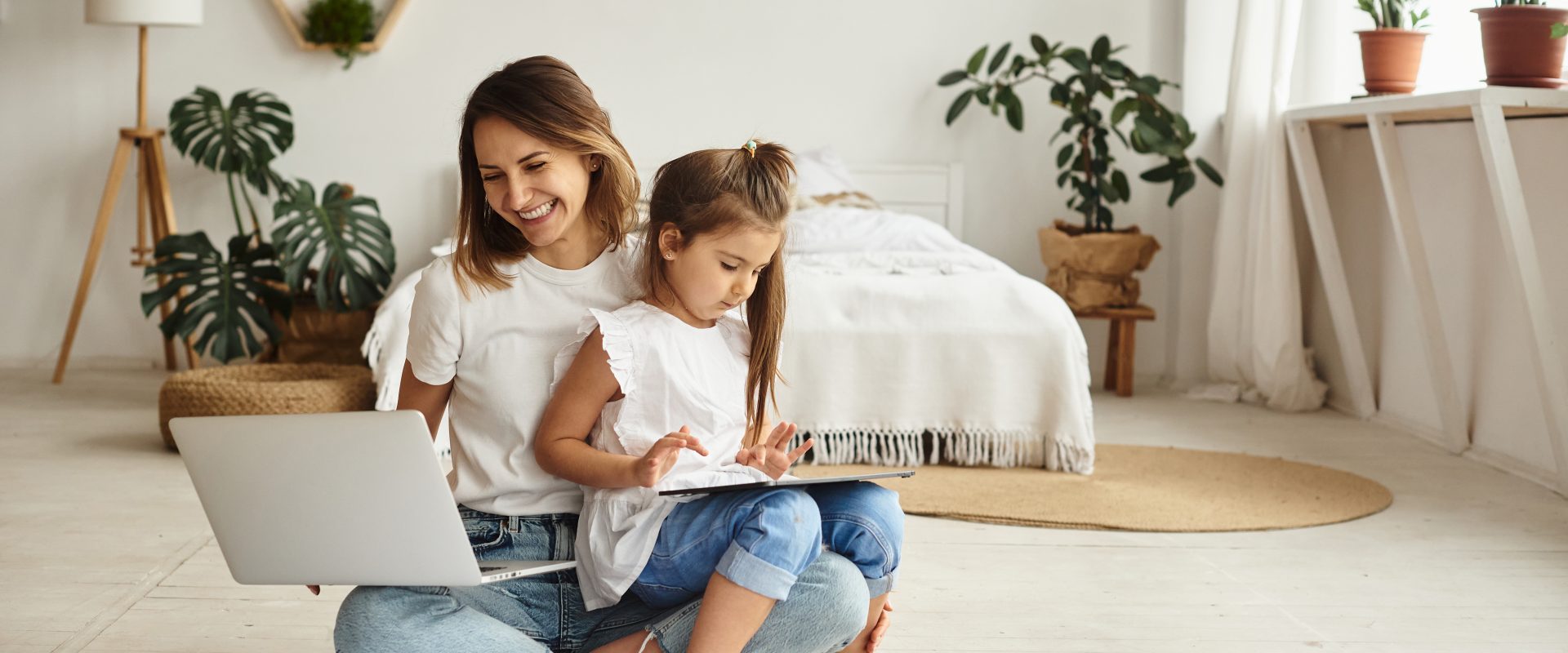 Współczesna mama - czy naprawdę musi być idealna? Mama pracująca w domu - siedzi na podłodze w sypialni, na jednym kolanie trzyma laptopa, na drugim kolanie siedzi jej córka o rysuje.