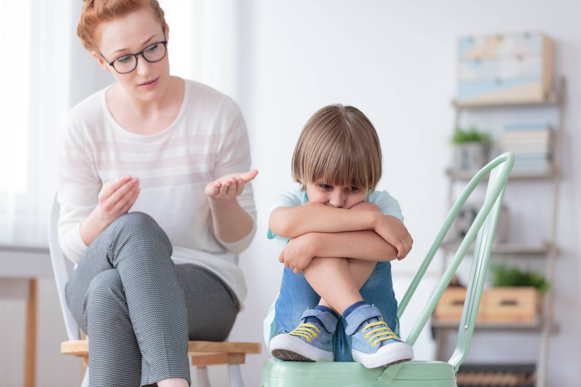 Wrażliwe dziecko. Obrażony chłopczyk siedzi z podkulonymi nogami na krześle, obok niego siedzi mama i tłumaczy mu coś.