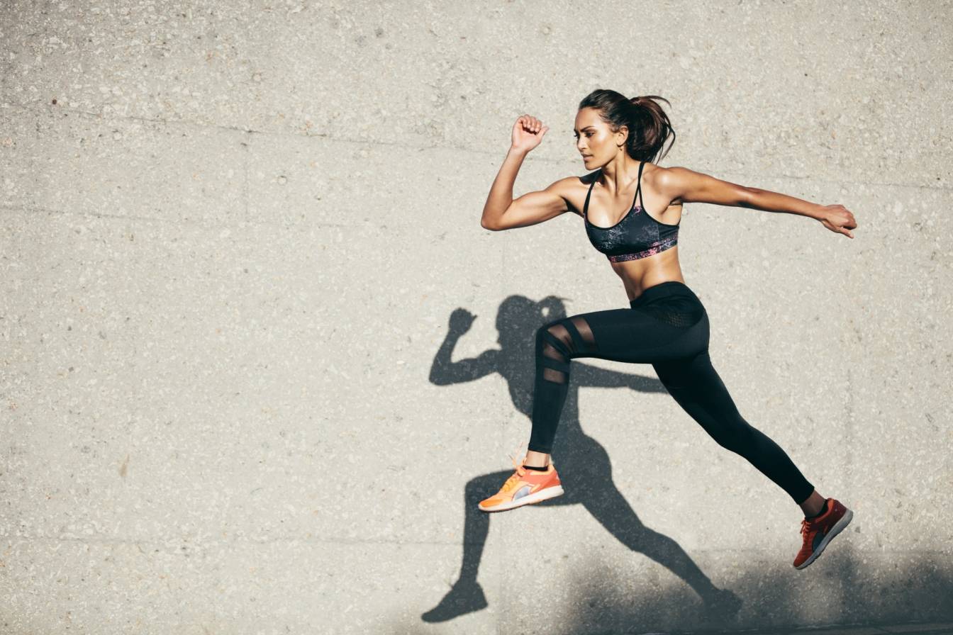 Sport amatorski/rekreacyjny a sport wyczynowy. Jakie są korzyści z uprawiania sportu, a jakie zagrożenia? Młoda wysportowana dziewczyna w stroju sportowym biegnie wzdłuż muru.