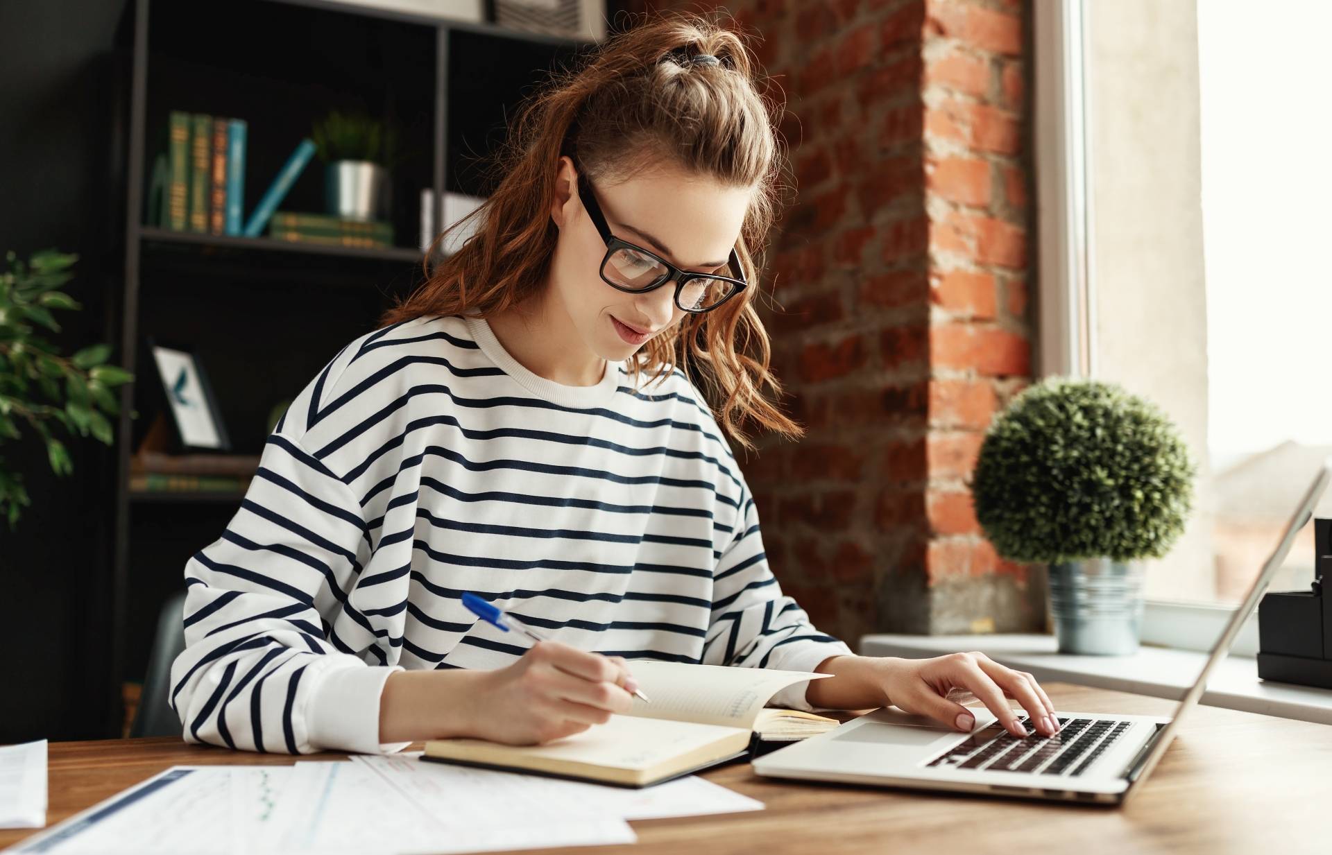 Work-life balance na home office. Młoda dziewczyna z pokolenia millenialsów w białej bluzce w czarne paski i okularach w rogowej oprawce siedzi przy laptopie i robi notatki w zeszycie.