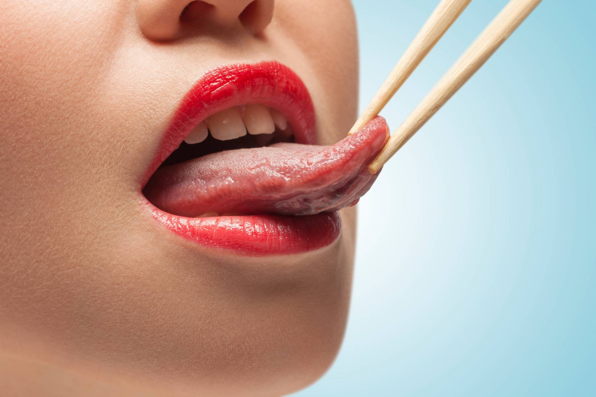 Jak wygląda zdrowy język? Zbliżenie na dolną część twarzy młodej kobiety, której język jest trzymany w patyczkach do sushi.