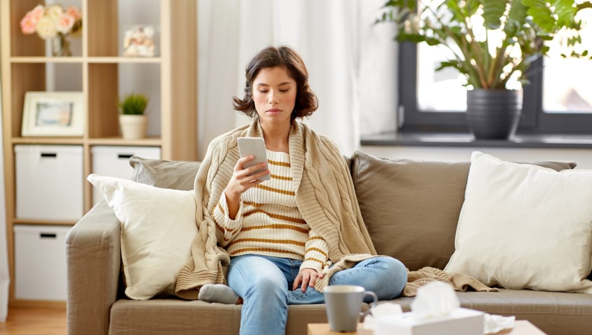 Post-COVID-19 Syndrome - zespół pocovidowy PCS - jakie są objawy i jak go leczyć? Zmęczona i chora kobieta zawinięta w koc siedzi na kanapie i przegląda smartfona.