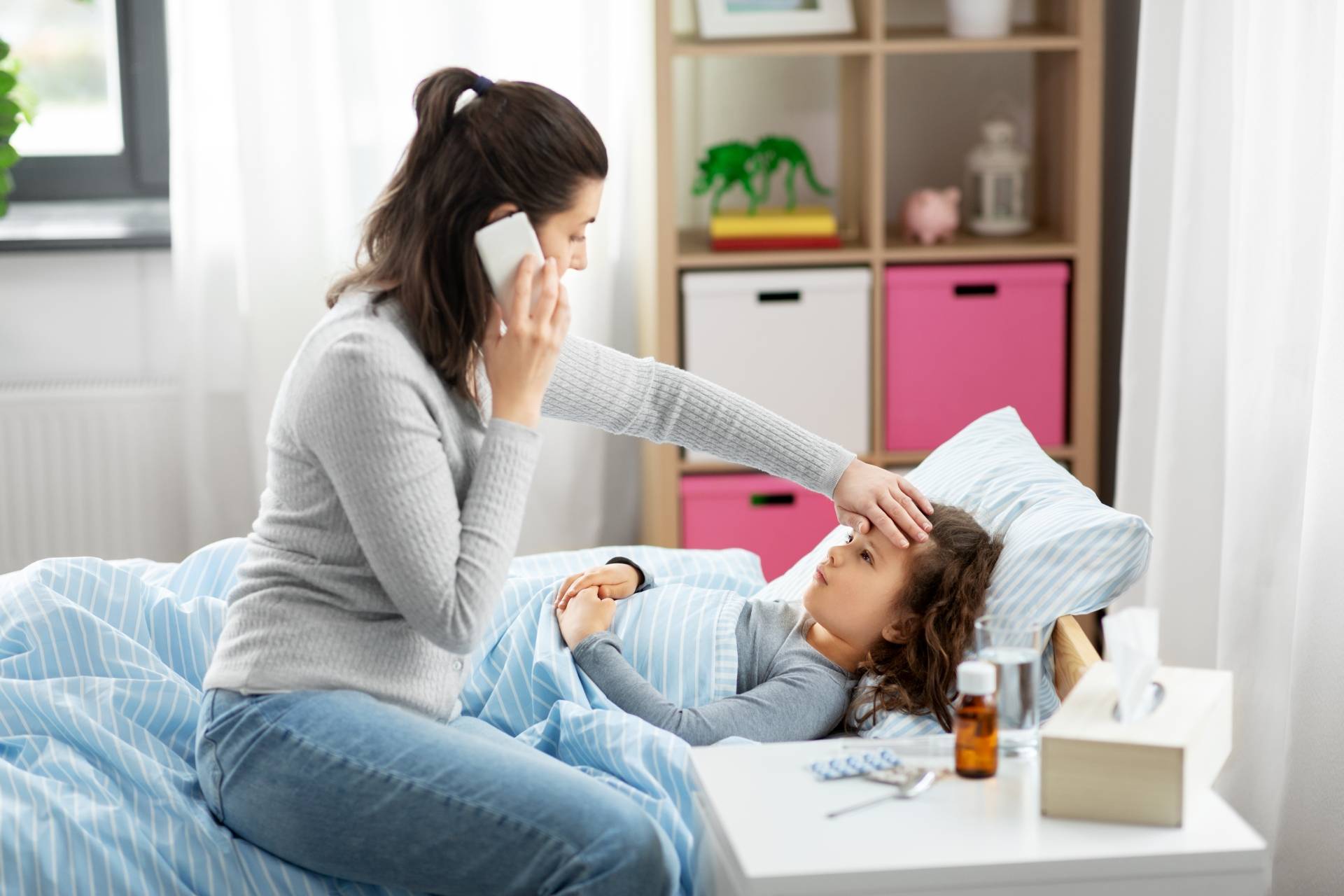 PIMS - zespół postcovidowy - jakie objawy powinny cię zaniepokoić? Mama mierzy temperaturę córce leżącej w łóżku i rozmawia przez telefon z lekarzem.