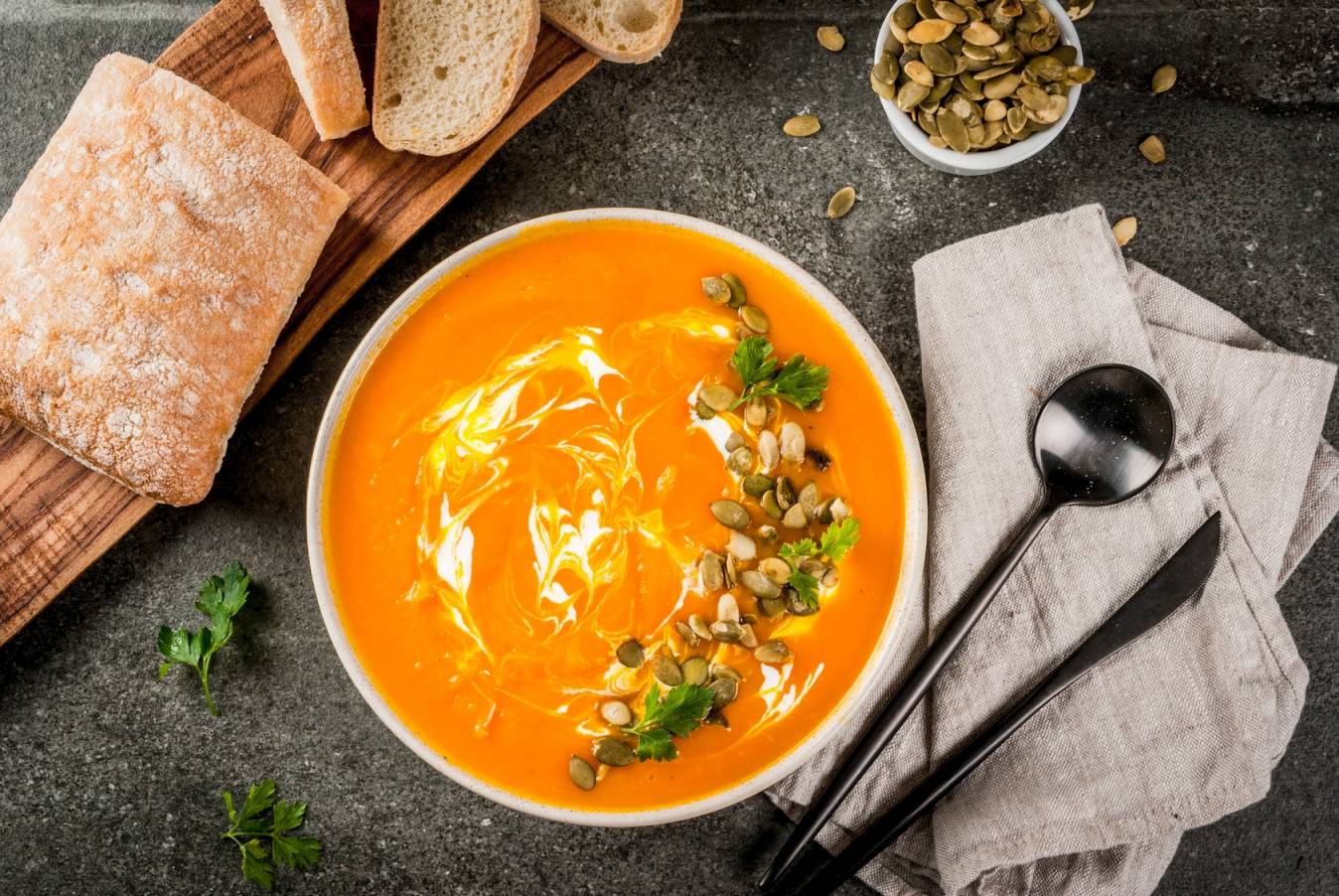 Dieta na przeziębienie wg medycyny chińskiej - co jeść , gdy dopadnie nas atak zimnego wiatru? Ujęcie z góry na miskę zupy dyniowej ze śmietaną, leżącą na szarym blacie, obok leżą deska z chlebem, ścierka i szczućce.